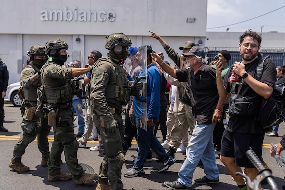 Heavily armed officers in tactical gear and face coverings stand in a line with clear riot shields as protesters confront them in the street. Several protesters gesture and record the interaction with their phones. The scene unfolds in front of a building marked "ambiance" on a bright day.